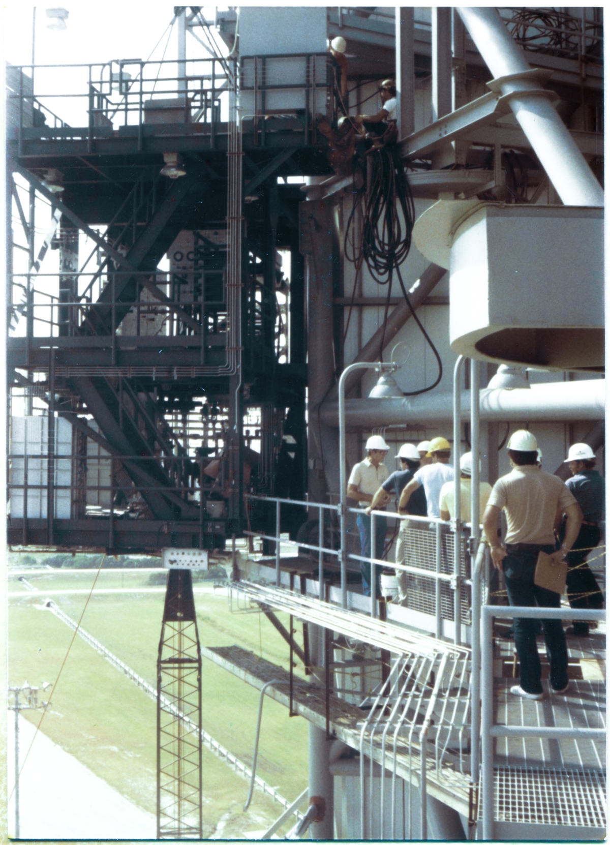 Image 095. Viewed from the portion of matching OMBUU Access Catwalk on FSS Side 1, used when the Rotating Service Structure at Space Shuttle Launch Complex 39-B, Kennedy Space Center, Florida, is in mated position, we see Wade Ivey, owner of Ivey Steel standing on the Access Catwalk, closest to the OMBUU, with his back toward it. Wade is discussing whatever interference that has interrupted the Lift and subsequent Connection with Contractor and Government personnel, and without doubt, he is explaining to everyone how he intends to proceed with the task as quickly as possible, and also without doubt, that task now includes some sort of torching-off or modifying one or more objects present on the RSS which will need to be removed in order for the OMBUU to be cleanly and properly bolted to the RSS. And whenever such interferences are encountered, you may rest assured that they belong to someone, and in ways we cannot know for this instance, the removal or modification will cause extra work, and extra expense, for the affected parties, in addition to causing extra work and extra expense for Ivey Steel, and Wade being Wade, my money is going with a guess that he's attempting to work with everyone involved, employing equitable give-and-take between parties, to smooth things along, and speed things along, with the least impact possible on the overall job. Wade was a man of his word, and he was known for being a man of his word, and this allowed him to organize and effect sudden changes in plan with other entities, verbally, on the spot, without waiting for paperwork to be created which would cover him (and them, too), and in this way the work would get done right away, with an absolute minimum of additional time and effort, and Wade would make good on such paper as would necessarily follow, formalizing the changed aspects of things, technically and contractually. Photo by James MacLaren.